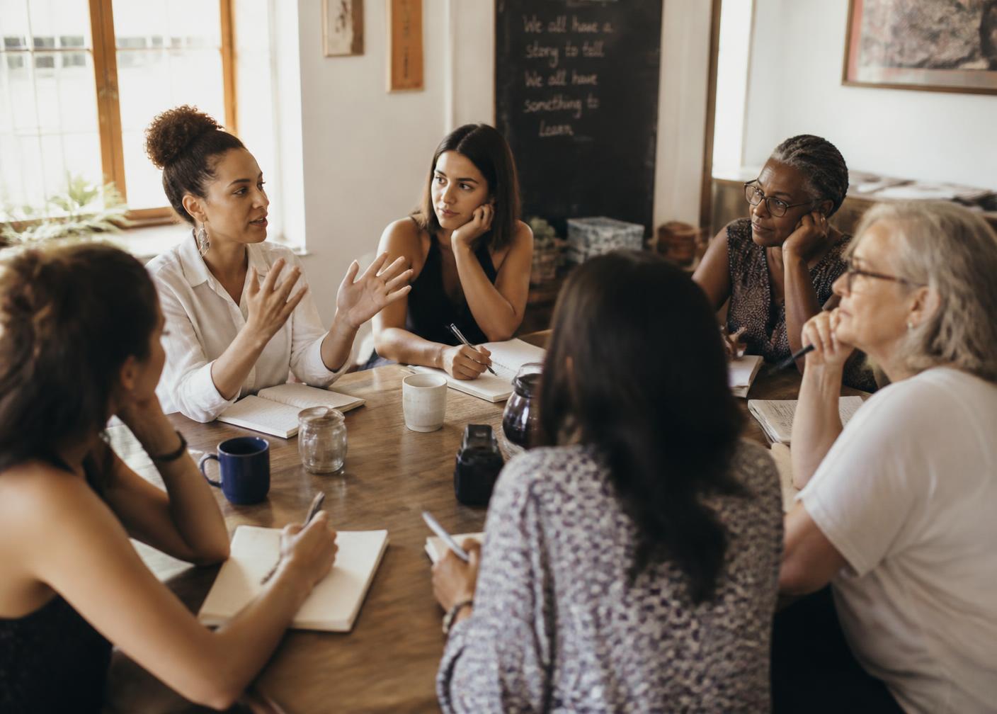 Women of many ages gathered around a table, listening and taking notes